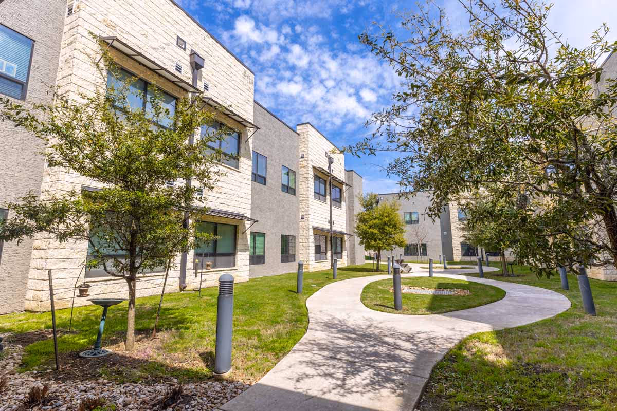 Curved concrete pathway surrounded by green grass and trees leading through a courtyard area between modern two-story buildings under a partly cloudy blue sky.