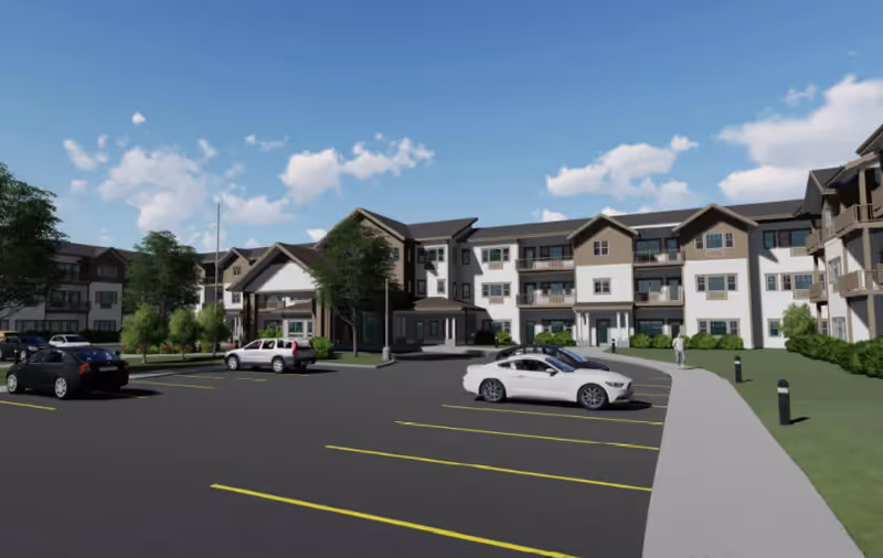 Exterior view of a large, multi-story senior living facility with a parking lot in front. Several cars are parked, and a few people are walking on the sidewalk near the building. The sky is clear with some clouds.
