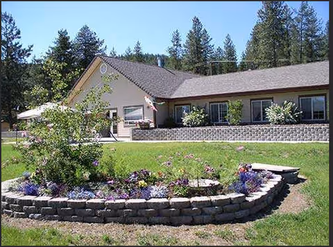 Single-story building with a sloped roof surrounded by trees and a well-maintained lawn featuring a raised flower bed with various colorful flowers in front.
