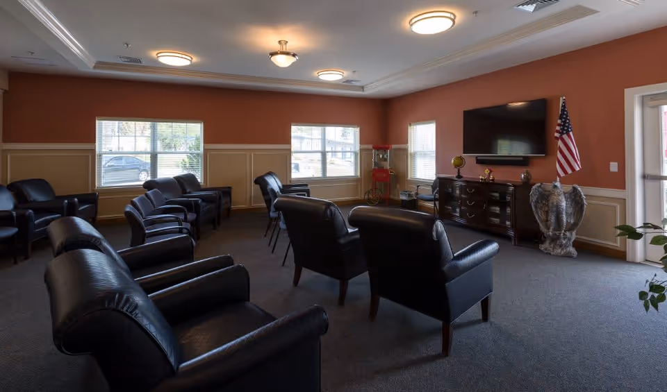 A common area with multiple black leather armchairs arranged in rows facing a wall-mounted flat screen TV. The room has three windows with blinds, a wooden cabinet under the TV, an American flag, and a decorative eagle statue. The walls are painted orange with beige wainscoting, and the ceiling has recessed lighting fixtures.