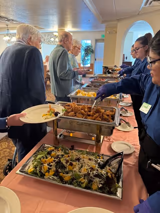 Several elderly people serving themselves food from a buffet table with trays of fried chicken, salad, and other dishes, assisted by staff wearing gloves and uniforms in a dining area with carpeted floor and large windows.