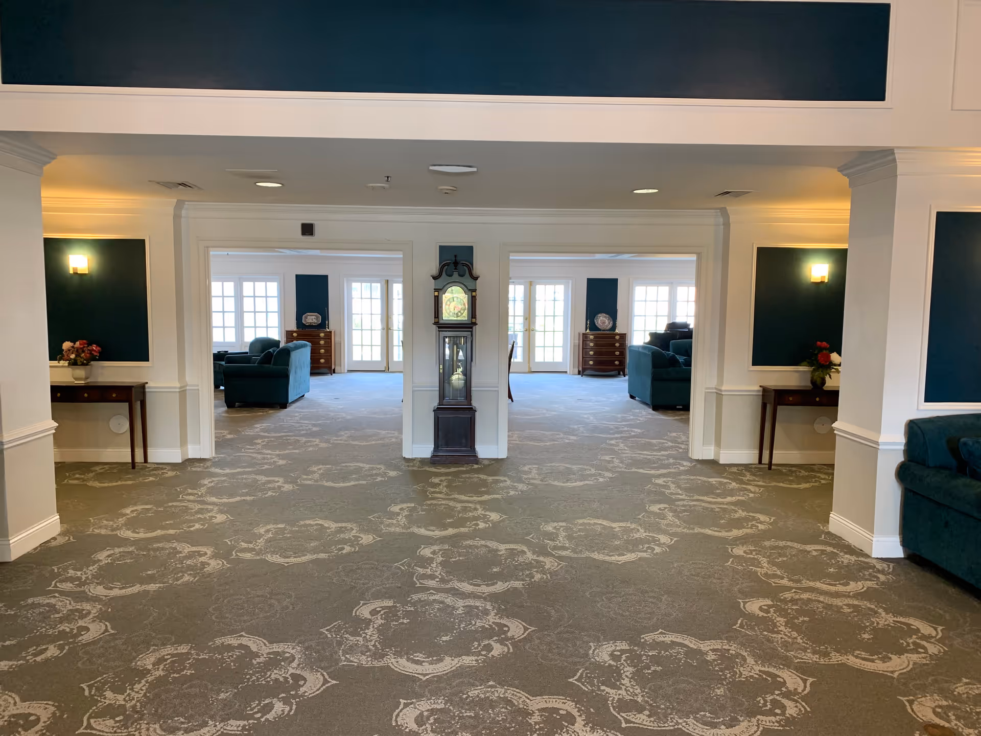 Interior view of a senior living facility lounge area with patterned carpet, white walls, and dark green accent panels. The space features a grandfather clock centered between two open doorways leading to rooms with teal sofas, wooden dressers, and large windows letting in natural light. Small tables with flower arrangements are placed against the walls.