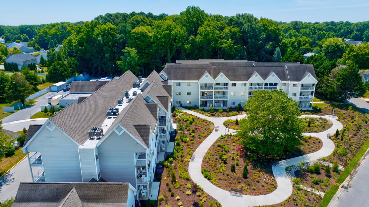 Aerial view of a senior living facility named Gull Creek showing multiple white residential buildings with gray roofs surrounding a landscaped garden area with winding concrete pathways and a large tree in the center. The background includes dense green trees and a clear blue sky.