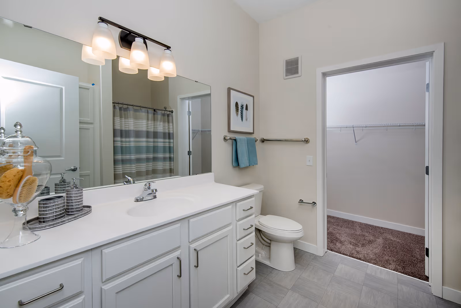 A clean and modern bathroom featuring a white vanity with a sink, a large mirror with four light fixtures above it, a toilet, and a doorway leading to a carpeted walk-in closet with wire shelving. The bathroom has light-colored walls and floor tiles, a striped shower curtain, and a towel rack with two blue towels hanging on it. Decorative items including a glass jar with bath sponges and a soap dispenser are placed on the vanity.