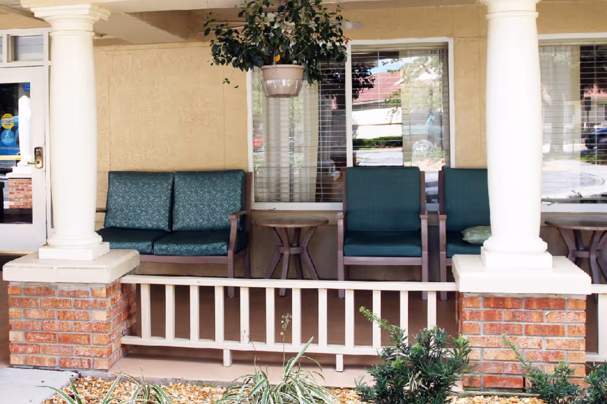 Covered porch area with green cushioned seating including a loveseat and two chairs, small wooden side tables, white columns with brick bases, a hanging potted plant, and windows with blinds behind the seating.