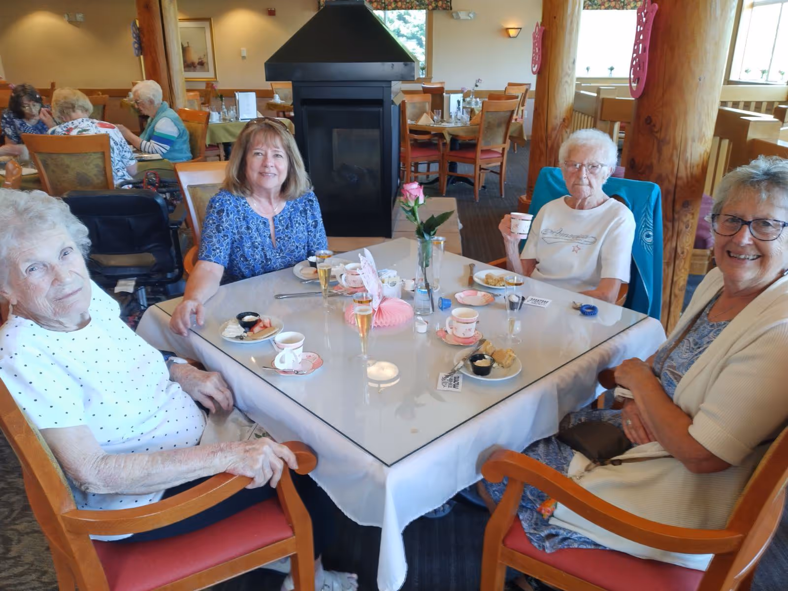 Four elderly women sitting around a square dining table with a white tablecloth in a dining room. They are enjoying tea and desserts, with cups, plates, and glasses on the table. The room has wooden pillars, large windows, and other tables with people in the background.