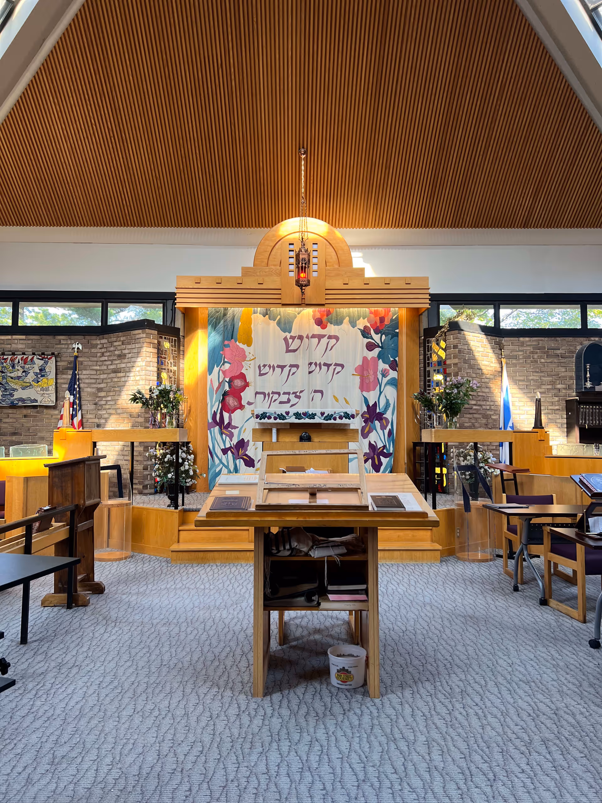 Interior view of a synagogue or prayer room with a wooden ark at the center, decorated with a colorful floral curtain featuring Hebrew text. The room has a high wooden slatted ceiling, carpeted floor, and various wooden furniture including a lectern and chairs. There are flags and floral arrangements on either side of the ark.