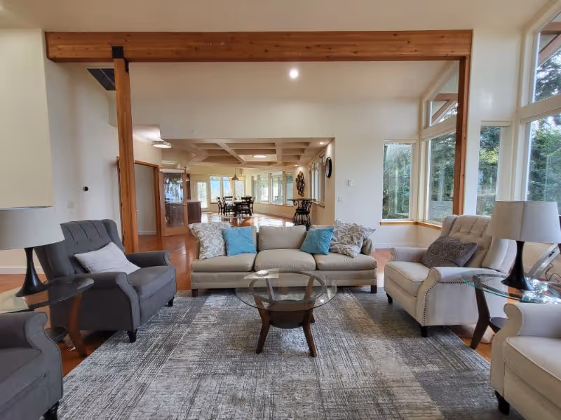A spacious living room with a beige sofa adorned with blue and patterned pillows, flanked by two gray armchairs and one beige armchair. There are glass-top side tables with lamps on either side of the seating area, and a round glass coffee table in the center. Large windows allow natural light to fill the room, and a wooden beam runs across the ceiling. In the background, a dining area with a table and chairs is visible.