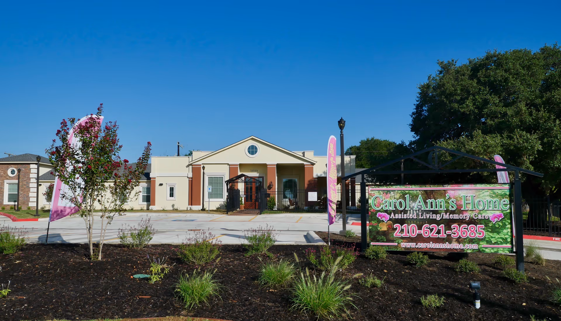 Front exterior view of Carol Ann's Home assisted living and memory care facility with a clear blue sky, landscaped garden area, and a sign displaying the facility's name, phone number, and website.