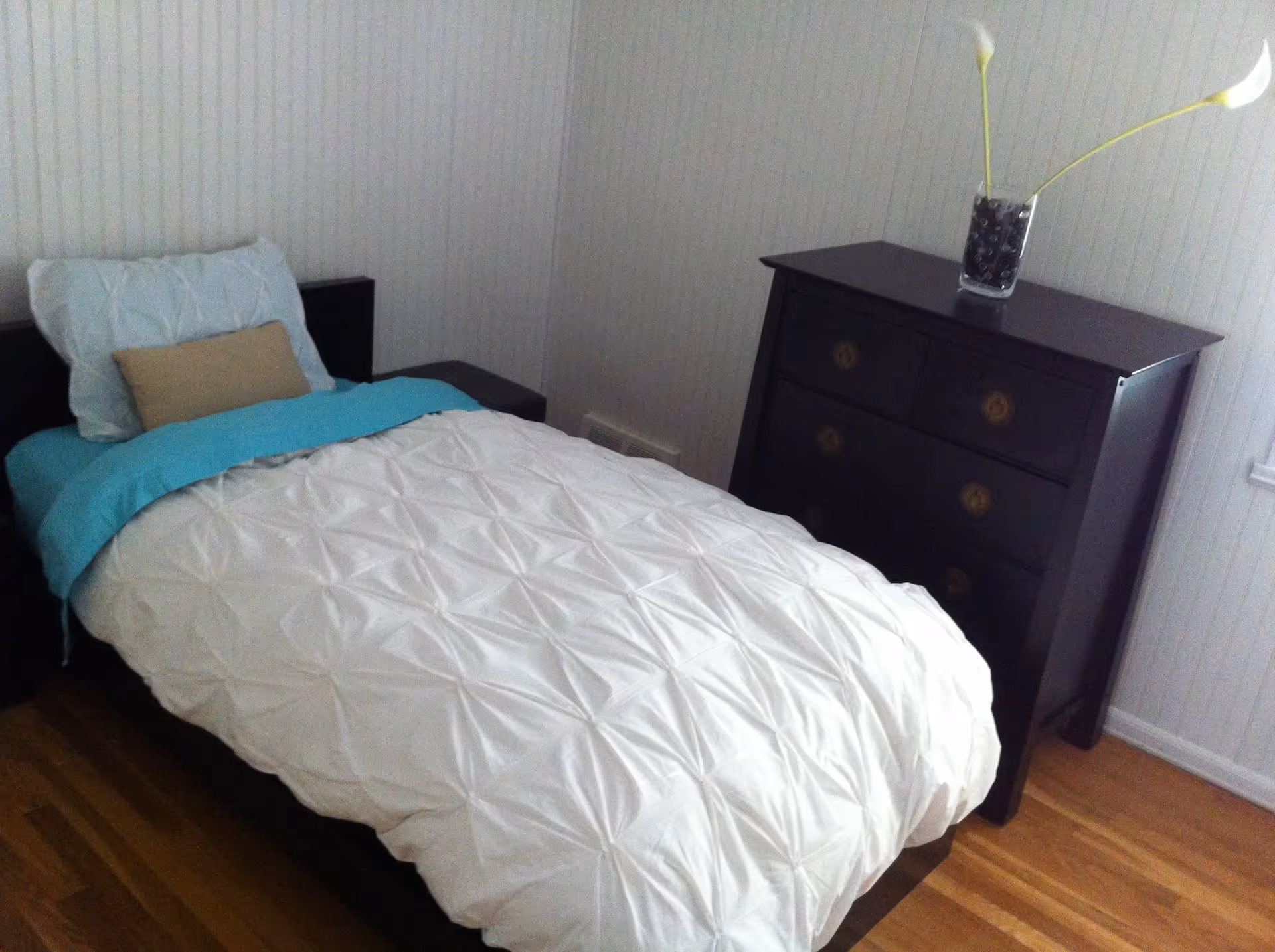 Single bed with a white textured duvet, teal throw and pillows next to a dark chest of drawers and hardwood floor in a small bedroom.