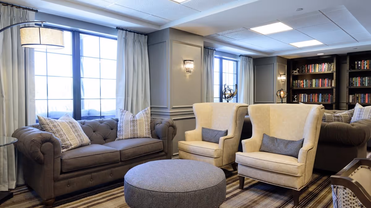 A cozy senior living facility lounge area with a dark brown tufted sofa adorned with patterned pillows, two beige wingback chairs with gray cushions, a round gray ottoman in the center, large windows with light curtains, and a bookshelf filled with books in the background.