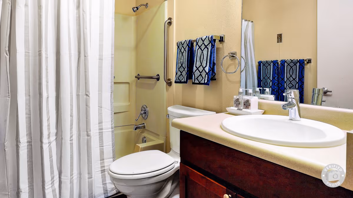 A clean bathroom with a white toilet, a sink with a countertop, a large mirror, and a shower area with a white and gray striped shower curtain. Two blue patterned towels hang on a towel rack above the toilet, and there are toiletries placed on the sink counter.