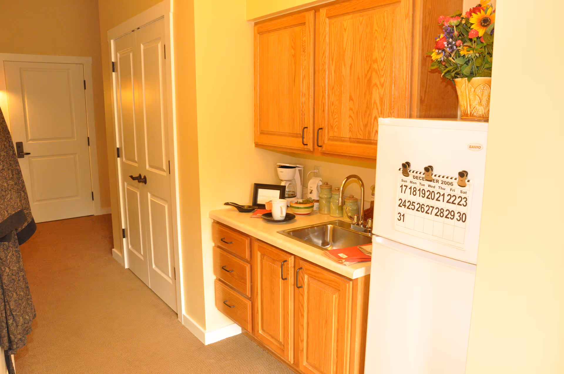A small kitchenette area with wooden cabinets, a countertop with a sink, a coffee maker, and some dishes. A white refrigerator with a calendar from December 2006 is visible on the right side. There is a hallway with closed white doors and a coat hanging on the left side.