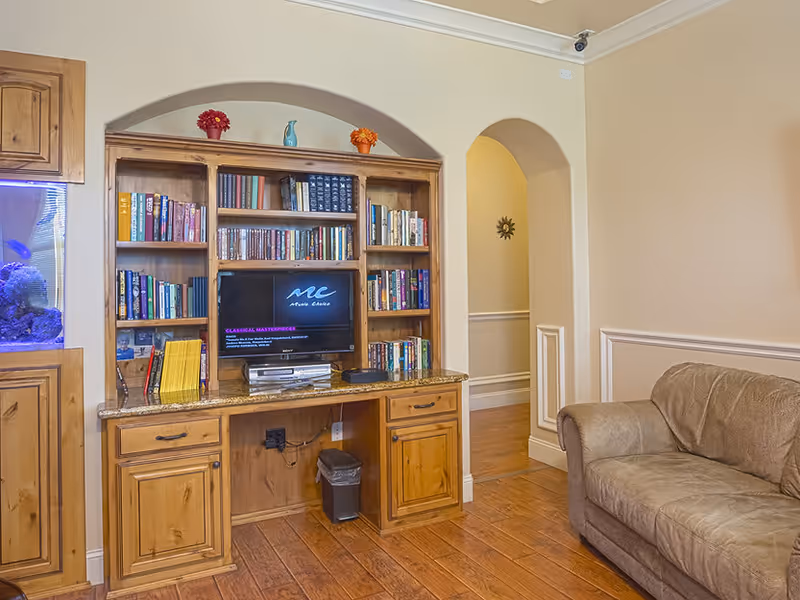 A cozy living room area featuring a wooden bookshelf with various books and a small TV displaying 'Music Choice'. To the left, there is a built-in aquarium with fish and coral. A beige couch is positioned on the right side, and an arched doorway leads to another room. The floor is wooden, and the walls are painted a light beige color.