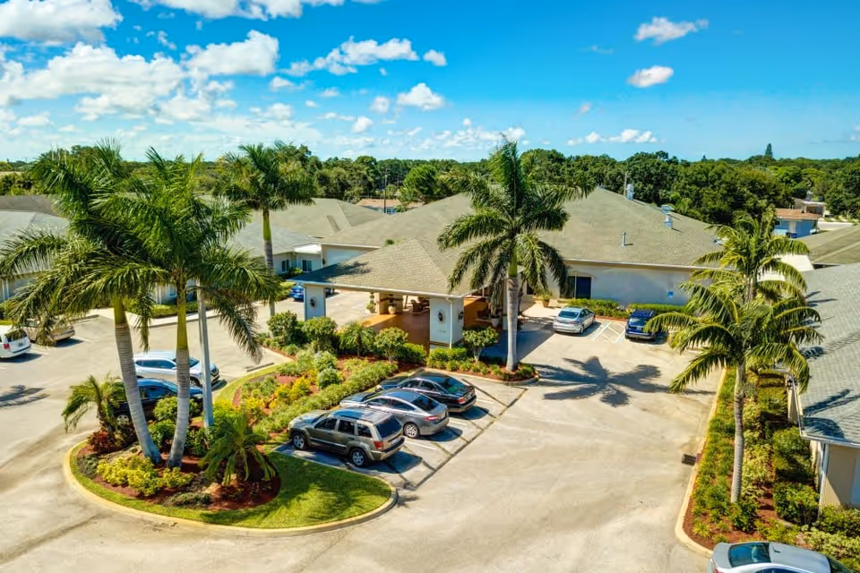 Aerial view of the entrance and parking area of Lynmore at Lawnwood facility, featuring several parked cars, palm trees, landscaped greenery, and a clear blue sky with scattered clouds.