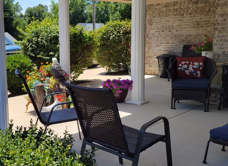 A covered outdoor patio area with black wicker chairs, one with a red floral cushion. There are potted flowers including purple and orange blooms, green bushes, and white columns supporting the roof. The background shows a driveway and more greenery under a clear sky.