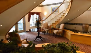 Lobby featuring a sweeping curved staircase, upholstered chairs around a central table, indoor plants and carpeting.
