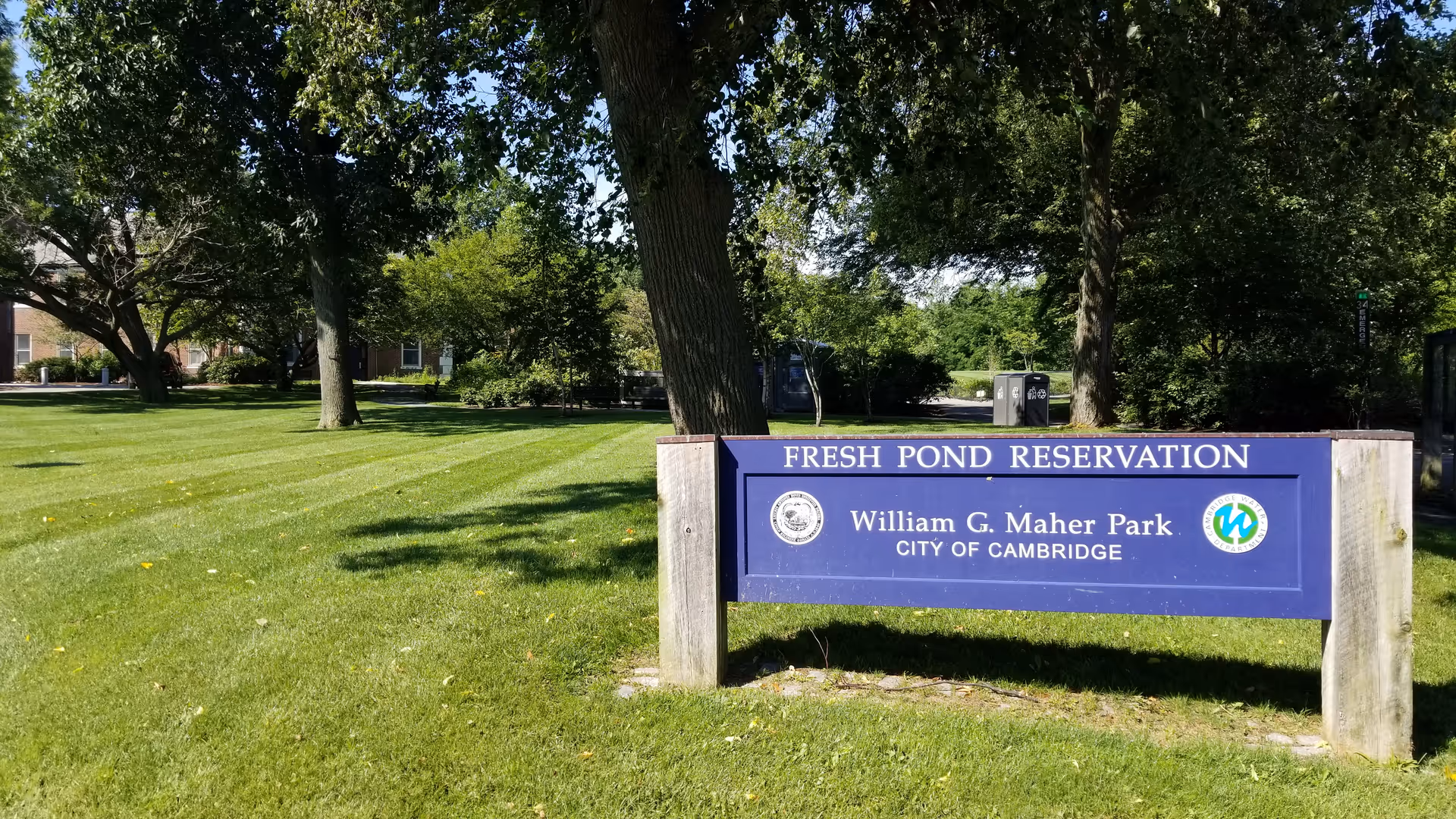 A green park area with neatly mowed grass and several large trees providing shade. In the foreground, there is a blue sign that reads 'FRESH POND RESERVATION William G. Maher Park CITY OF CAMBRIDGE'. The background shows more trees and a glimpse of a building partially visible on the left side.