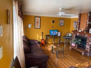 Cozy living room with mustard yellow walls, a dark brown leather sofa, a glass coffee table, a guitar leaning against the wall, a small TV on a stand, a globe on a side table, a brick fireplace with plants on the mantel, and hardwood floors.