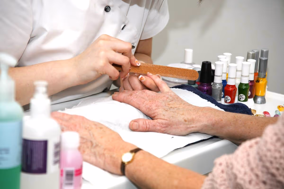A close-up view of a person filing the nails of an elderly person's hand during a manicure session. Various nail polish bottles and manicure tools are visible on the table.