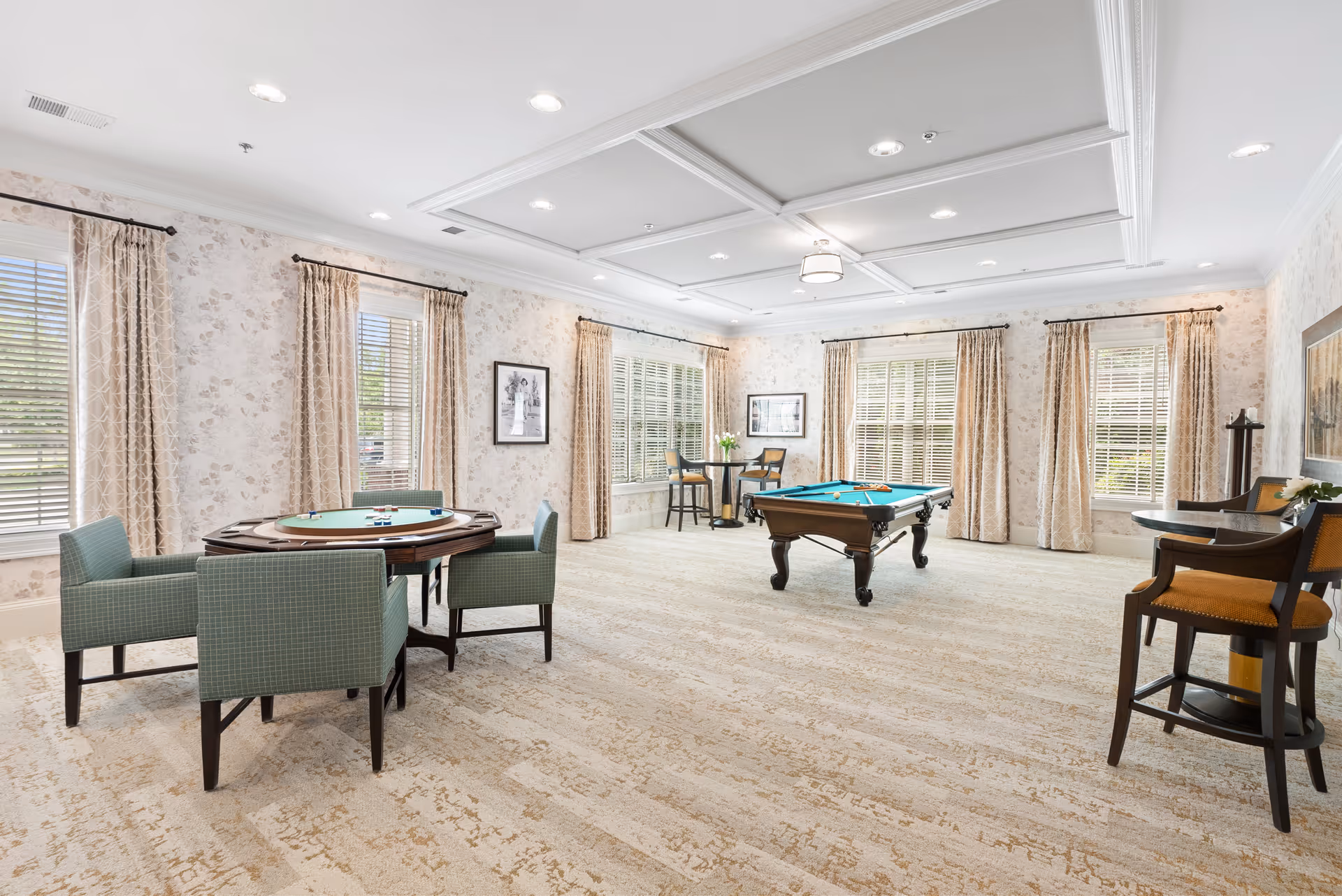 A spacious, well-lit game room with large windows covered by beige curtains. The room features a pool table with cues and balls, a round card table with four green upholstered chairs, and two small high tables with chairs. The walls have light floral wallpaper and framed black-and-white photographs. The ceiling has recessed lighting and decorative molding.
