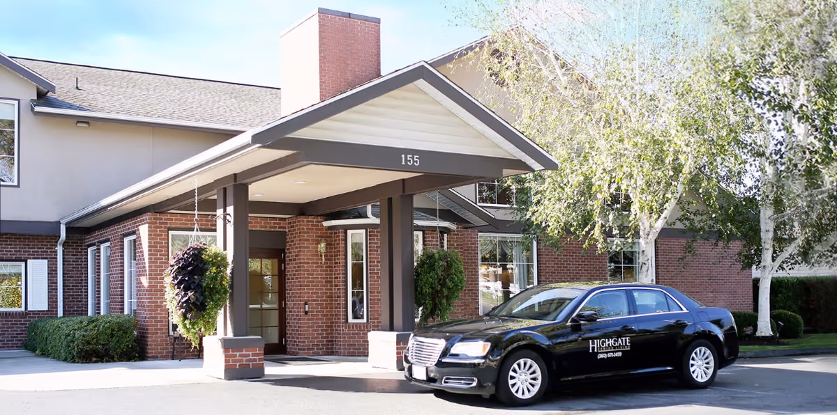 Exterior view of a senior living facility entrance with a covered drop-off area supported by pillars. A black car with the Highgate Senior Living logo is parked in front. The building is made of red brick with white trim and surrounded by trees and shrubs.