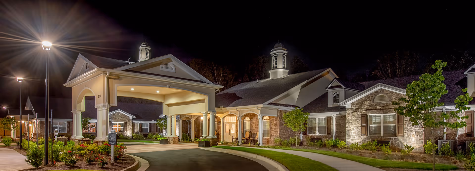 Night view of Benton House of Decatur senior living facility entrance with well-lit covered driveway, stone and brick exterior, landscaped greenery, and street lamps.
