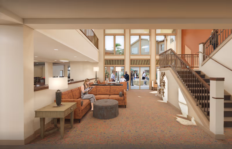 A spacious senior living facility common area with a large orange sectional sofa, a round ottoman, and a wooden side table with a lamp. A woman is sitting on the sofa reading a magazine. The area features high ceilings with large windows letting in natural light, and a staircase with wooden railings on the right side. Several people are visible in the background near the entrance and seating area.