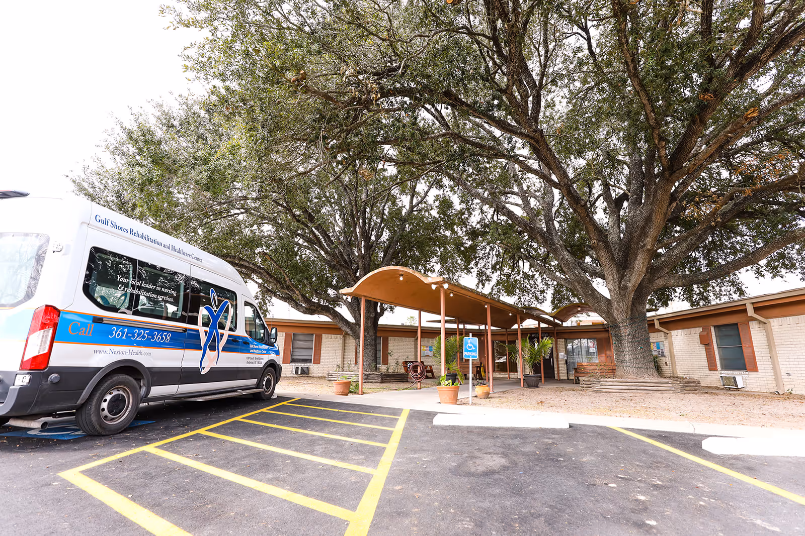 Exterior view of Gulf Shores Rehabilitation and Healthcare Center showing a white and blue facility van parked in a handicapped parking space near the entrance. The entrance has a covered walkway with large trees providing shade. The building is single-story with light-colored brick walls and several windows.