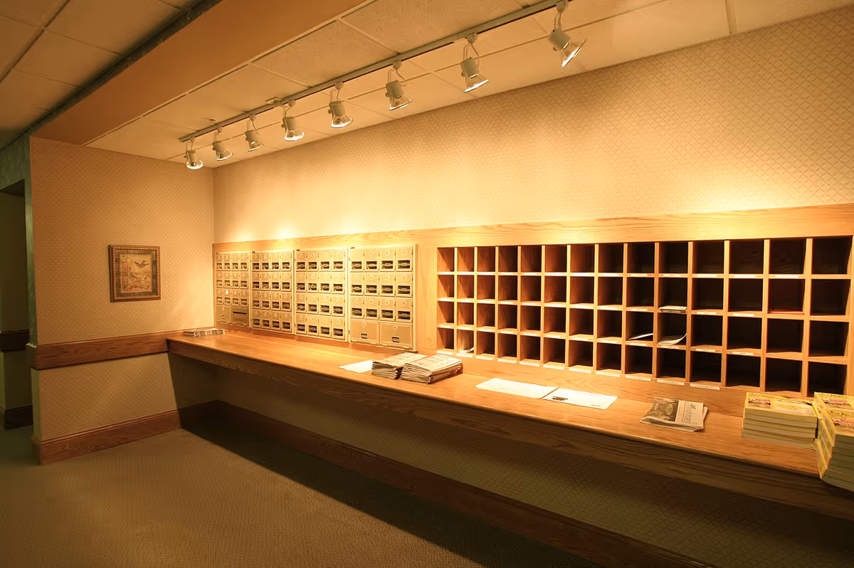 Interior view of a mailroom area with multiple mailboxes and wooden cubbyholes on the wall. There are stacks of newspapers and magazines placed on a wooden counter beneath the mailboxes. The room is warmly lit with ceiling track lights.