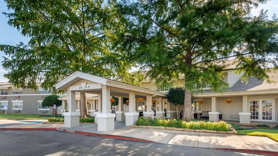 Covered front entrance and porte-cochere of a senior living building surrounded by large trees and landscaped planters.