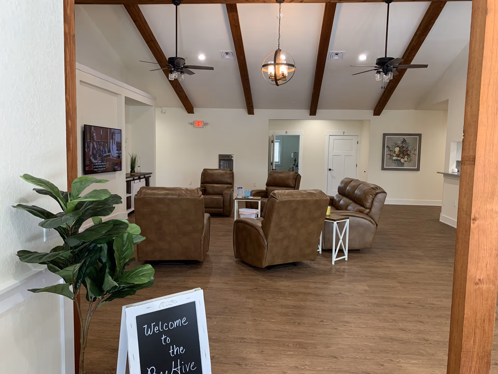 A spacious living room area with wooden beams on the ceiling, four brown leather recliners arranged around a small table, a wall-mounted TV on the left, a potted plant, and a chalkboard sign that reads 'Welcome to the BeeHive'. The room has light-colored walls, wood flooring, and a framed floral painting on the far wall.