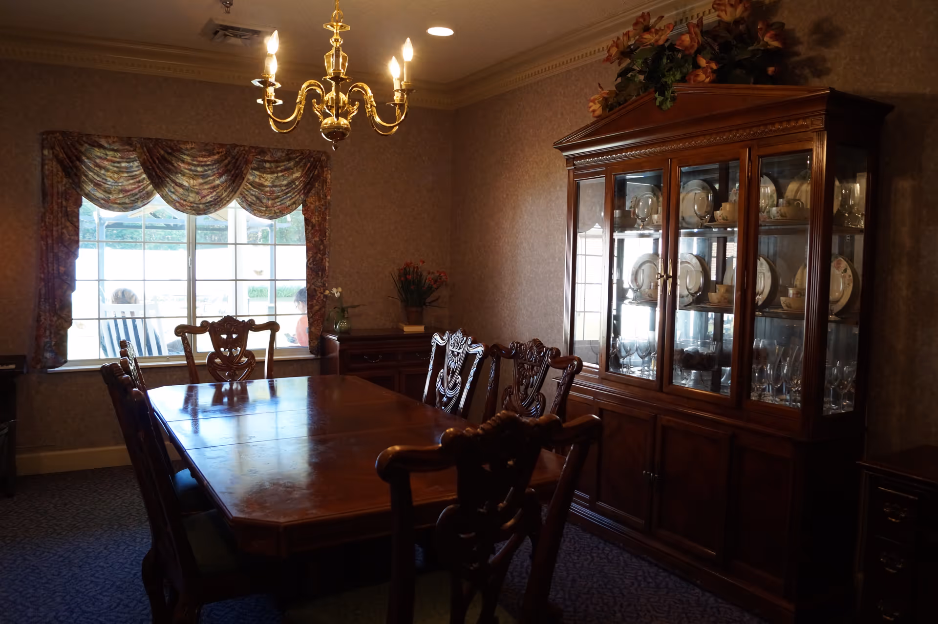 A traditional dining room with a wooden dining table surrounded by six ornate wooden chairs. A large wooden china cabinet filled with plates, glasses, and cups is against the right wall. A window with patterned curtains lets in natural light, and a brass chandelier hangs from the ceiling above the table. The room has wallpapered walls and carpeted flooring.