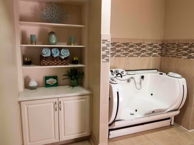 A bathroom area featuring a white walk-in bathtub with built-in controls and a tiled wall with a mosaic accent strip. To the left, there is a built-in shelving unit with decorative items including rolled towels, candles, a small plant, and a sign that reads 'EVERYDAY'.