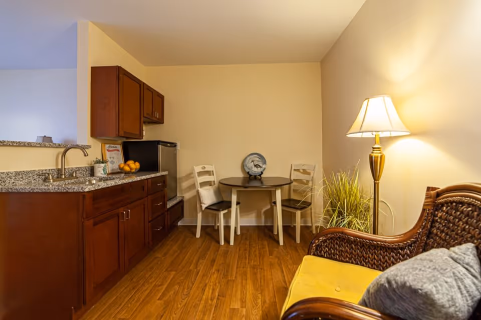 A cozy interior space featuring a small kitchenette with wooden cabinets, a granite countertop, and a stainless steel sink. Next to the kitchenette is a small round dining table with two white chairs, one with a cushion. A decorative plate is displayed on the table. To the right, there is a wicker armchair with a yellow cushion and a gray pillow, a tall floor lamp with a white shade, and a potted plant. The room has wooden flooring and beige walls.