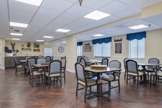A spacious dining room with multiple tables and chairs arranged neatly. The room has large windows with blue valances, light yellow walls, and a wood-patterned floor. There are decorative items on the walls, including framed pictures and signs that say Baseball and Football. The ceiling has recessed lighting panels.
