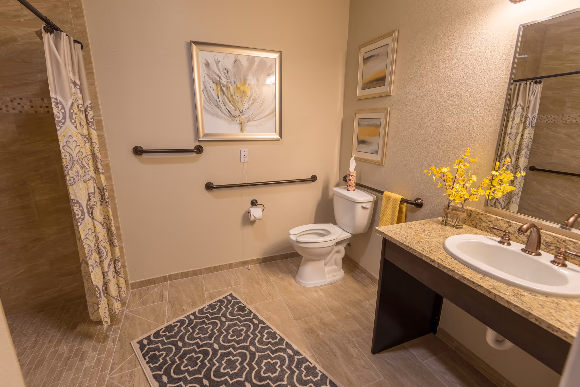 A clean and well-lit bathroom with beige tiled floors and walls. There is a toilet with a yellow towel hanging on a grab bar beside it, a granite countertop with a sink and bronze faucet, and a vase with yellow flowers on the counter. The shower area has a patterned curtain and tiled walls. The bathroom also features framed artwork on the walls and a patterned rug on the floor.