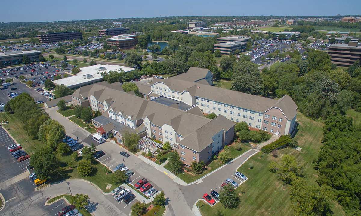Aerial view of Silvercrest at Deer Creek Senior Living facility showing a large multi-wing building surrounded by parking lots, trees, and green lawns. The building has a mix of brick and light-colored siding with multiple windows and a main entrance with a covered drop-off area. The surrounding area includes office buildings, parking lots, and a small pond in the distance under a clear sky.