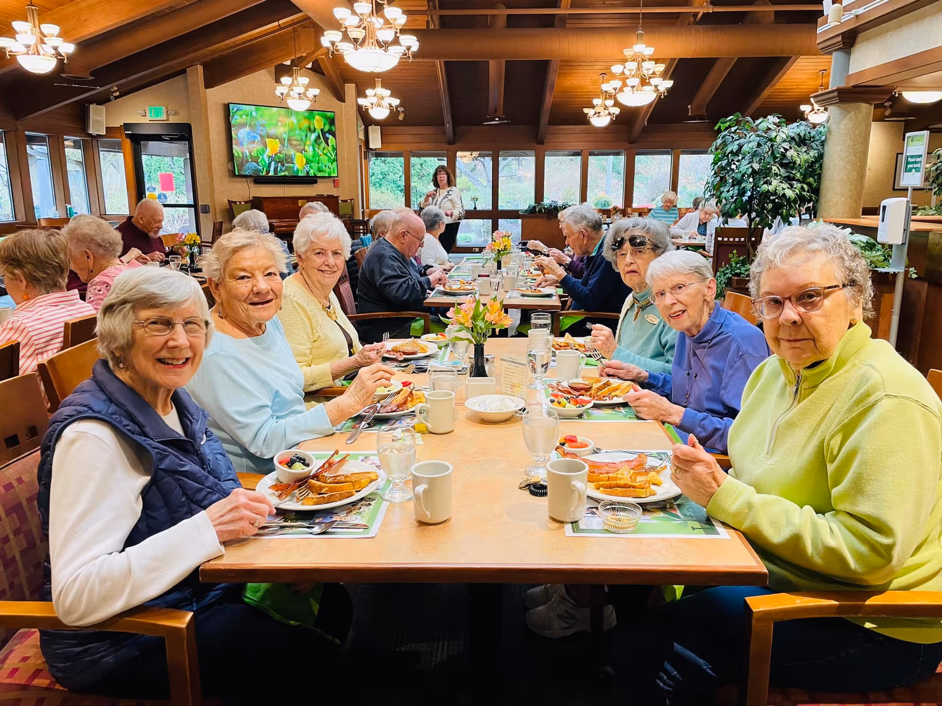A group of elderly people sitting around a long dining table in a well-lit room with large windows and wooden beams on the ceiling. They are enjoying a meal together, with plates of food, cups, and glasses of water in front of them. The atmosphere appears warm and social, with some people smiling at the camera. There is a television mounted on the wall in the background showing a nature scene, and a woman standing near the back of the room.