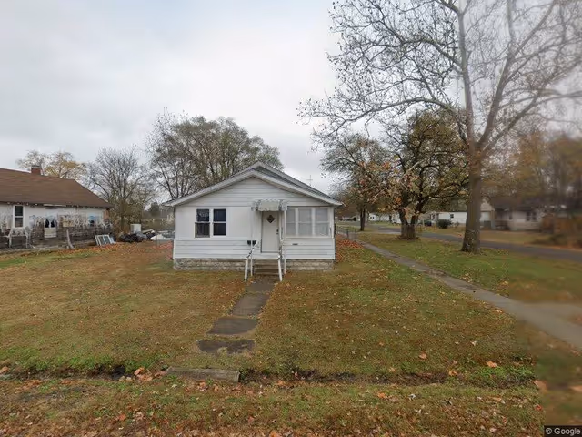 Small white single-story house with a front porch and steps leading to the door, situated on a grassy lot with a sidewalk and trees nearby under a cloudy sky.