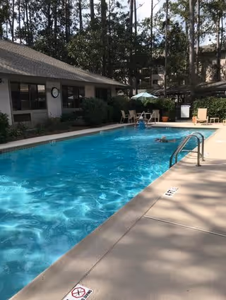 Outdoor swimming pool at a senior living facility with clear blue water, surrounded by a concrete deck with lounge chairs and umbrellas. Trees and buildings are visible in the background.