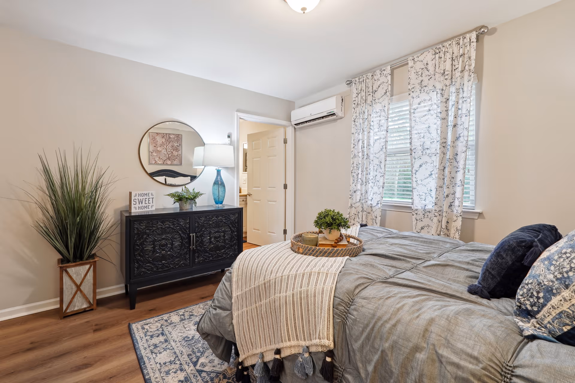 A cozy bedroom with a neatly made bed featuring gray bedding and decorative pillows. A woven tray with a small plant and candles sits on the bed. The room has light-colored walls, a window with patterned curtains, a black ornate dresser with a round mirror above it, a blue glass lamp, and a potted plant in a wooden planter. The door to an adjoining room is open.