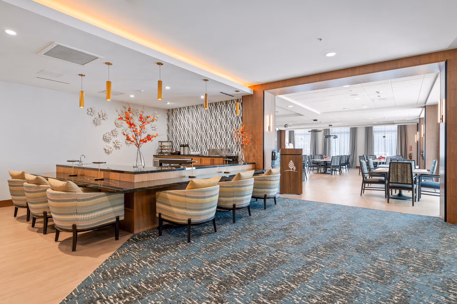 A modern dining area in a senior living facility featuring a long counter with cushioned chairs, decorative vases with orange flowers, pendant lighting, and a patterned accent wall. Beyond the counter, there is a spacious dining room with multiple tables and chairs, large windows with sheer curtains, and soft lighting.