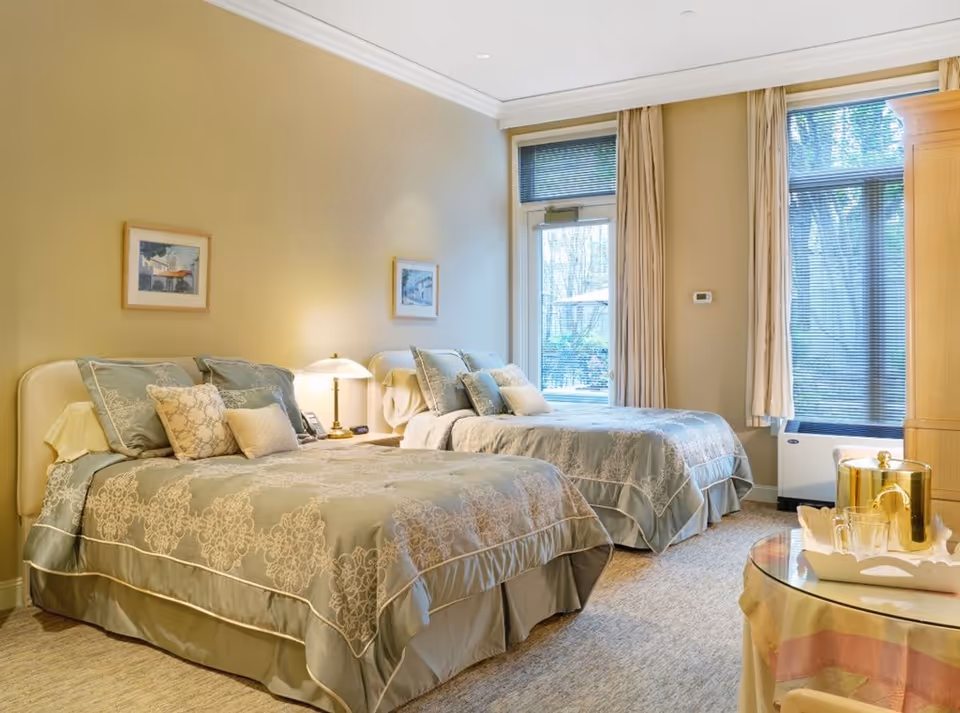 Bright, well-appointed bedroom with two twin beds, decorative bedding, a bedside lamp and large windows with curtains.