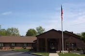 Exterior view of a single-story brick building with a peaked roof and an American flag on a flagpole in front, under a clear blue sky.