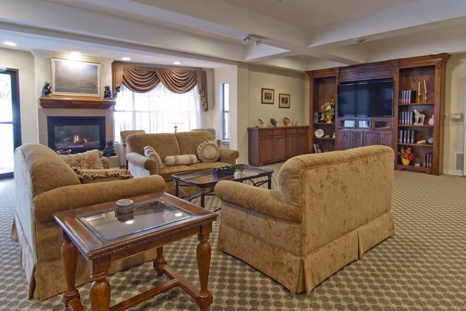 A cozy living room with beige upholstered sofas arranged around a glass coffee table. There is a fireplace with a painting above it on the left side, and a large wooden entertainment center with a flat-screen TV and decorative items on the right. The room has patterned carpet, a window with draped curtains, and warm lighting.