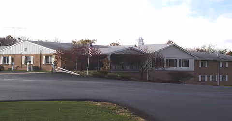 Front exterior of the Omni West Assisted Living building with a paved driveway, American flag, and small cupola.
