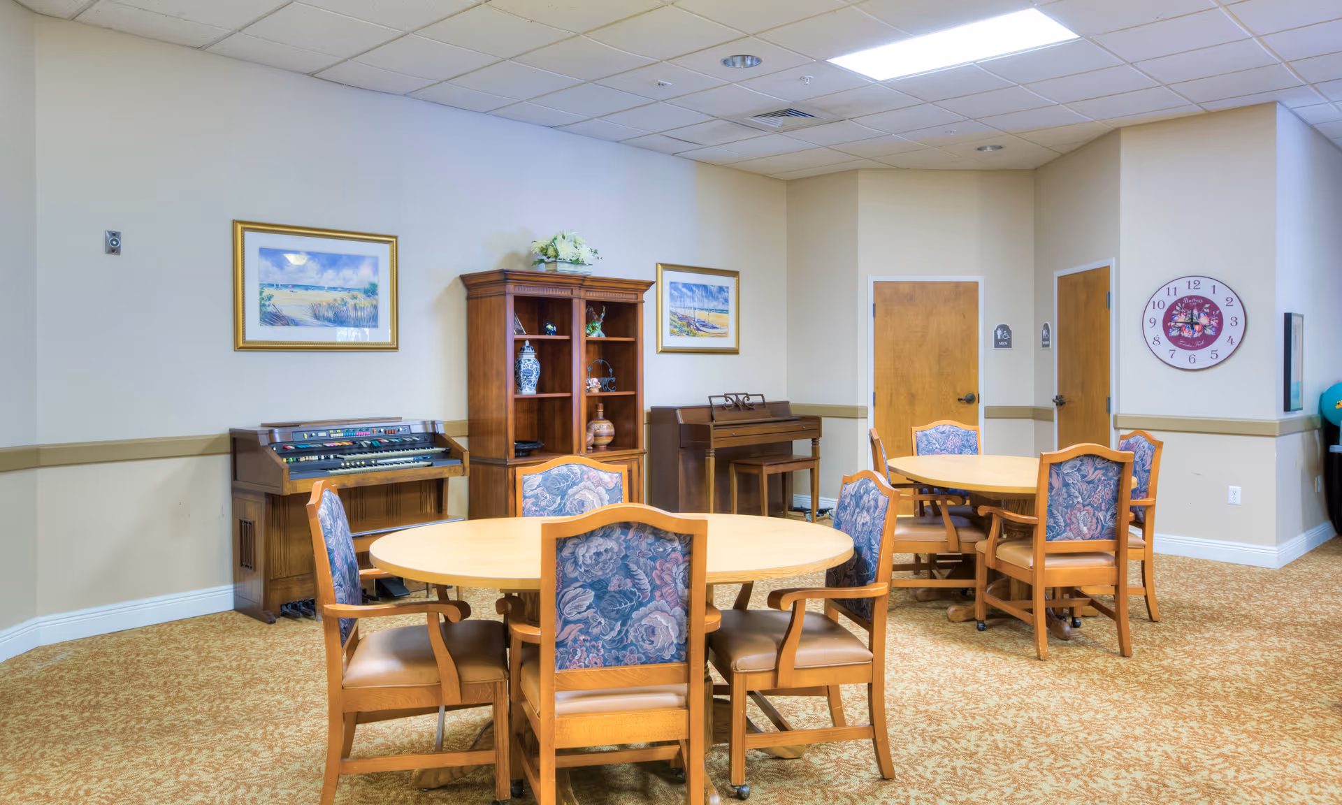 A bright common/dining room with round wooden tables and upholstered chairs, a piano and shelving against a beige wall.