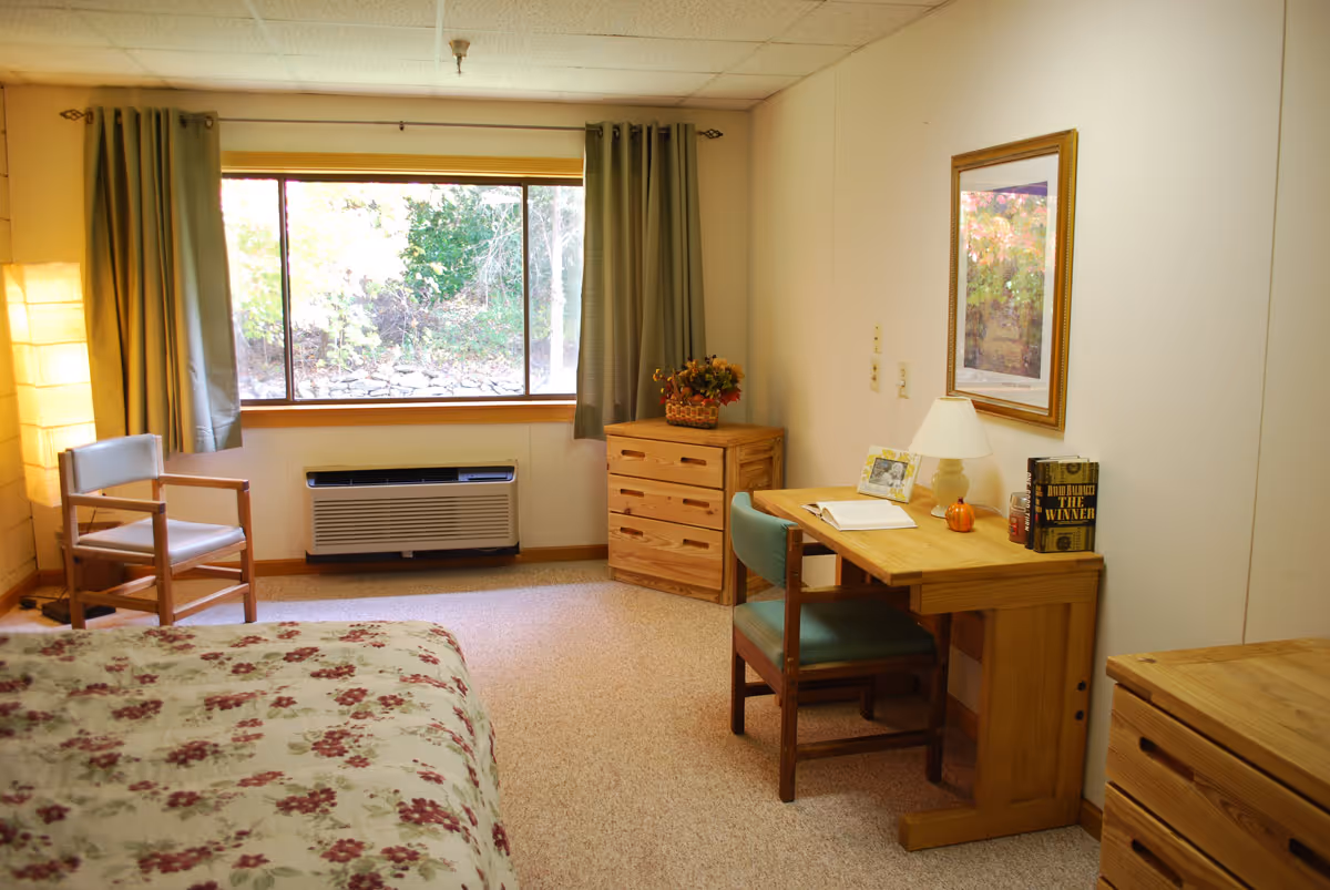 A cozy bedroom with a floral bedspread, a wooden chair, a wooden desk with a chair, a lamp, books, and a framed picture on the wall. A large window with green curtains lets in natural light, and there are wooden dressers in the room.