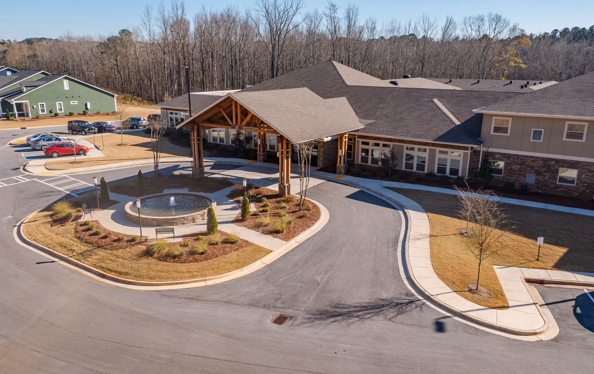 Aerial view of the entrance to Oaks at Eagle's Landing senior living facility showing a covered drop-off area with wooden beams, a circular driveway, landscaped garden with a small fountain, and parked cars. The building is surrounded by trees and has a mix of stone and siding exterior.